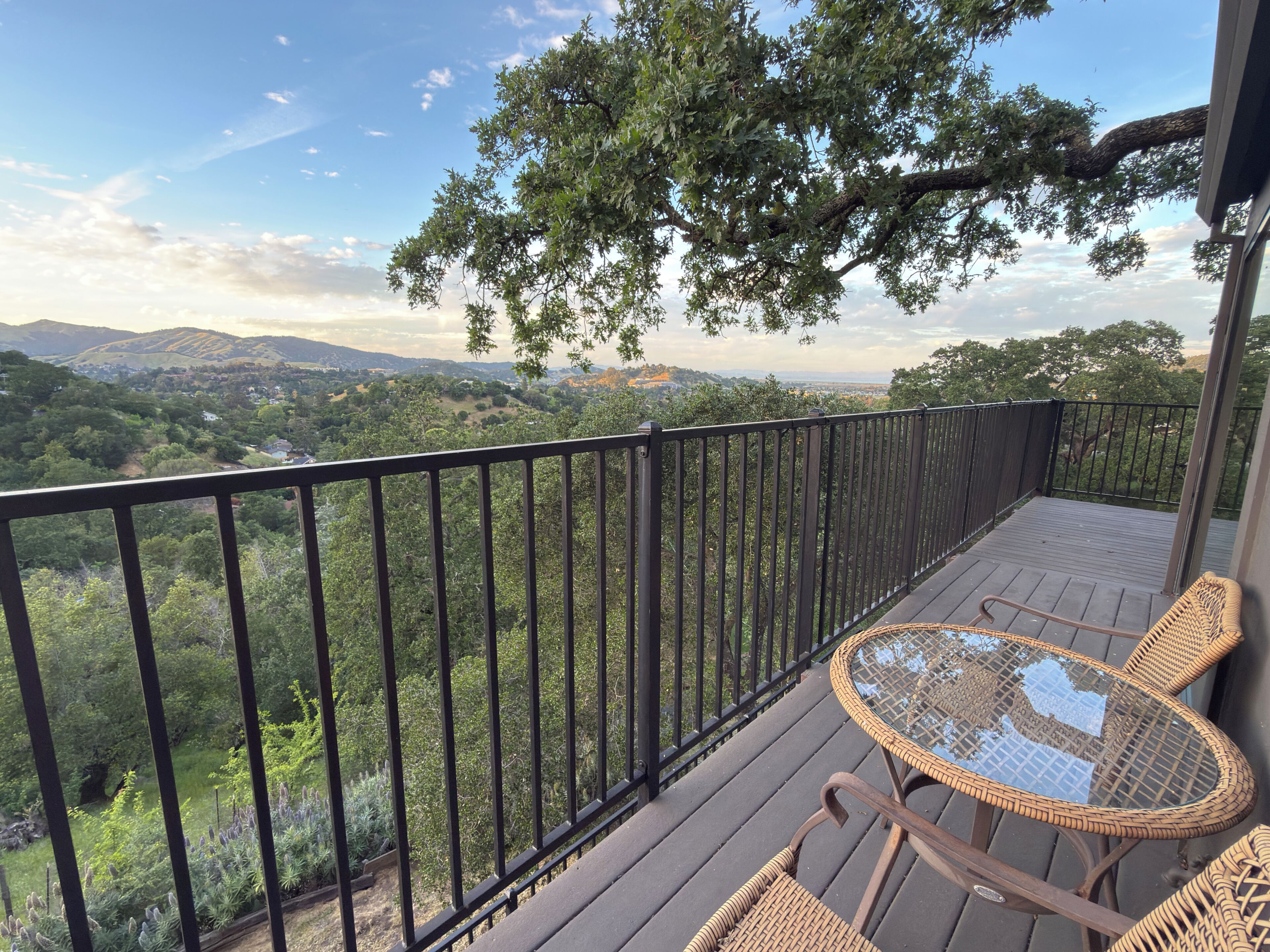 A balcony with a wicker table and chairs, overlooking a scenic landscape with trees and hills.