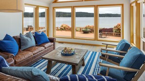 A cozy room with a brown leather sofa, blue and gray cushions, a wooden coffee table, and a striped rug. Large windows offer a view of a lakeside deck with outdoor furniture and flowers. - Spacious Beach Front Home on Hood Canal w/ Kayaks! (Belfair)