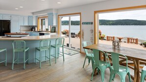 A modern kitchen with a large island, a dining area with chairs, and a view of a lake through large windows. - Spacious Beach Front Home on Hood Canal w/ Kayaks! (Belfair)