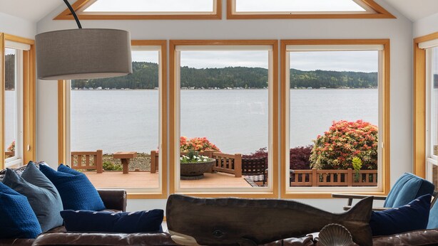 A room with a large bay window overlooking a lake, a wooden deck with outdoor furniture, and a view of a forested hill. - Spacious Beach Front Home on Hood Canal w/ Kayaks! (Belfair)