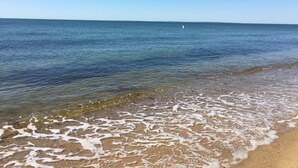 Une plage à l’eau claire, un rivage sablonneux et un voilier à l’horizon.