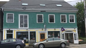 A two-story building with a green facade and a yellow storefront. There are two cars parked in front, and a sign indicating '122 Cottage' on the ground floor. - Recently Renovated In-Town Bar Harbor 4 Bed 3 Bth Apartment with Private Parking (Bar Harbor)