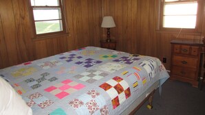 A bedroom with a large bed, wooden walls, two windows with white curtains, a nightstand with a lamp, and a dresser. - Come stay in our 2bd cabin on Table Rock Lake close to Silver Dollar City! (Branson)