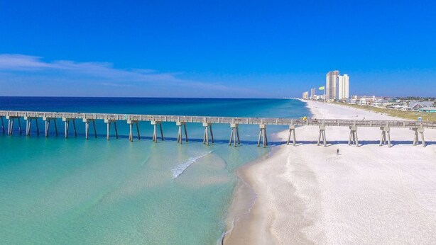 Una playa con un muelle largo que se adentra en el océano, una orilla arenosa y edificios al fondo.