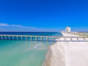 A beach with a long pier extending into the ocean, a sandy shore, and buildings in the background.