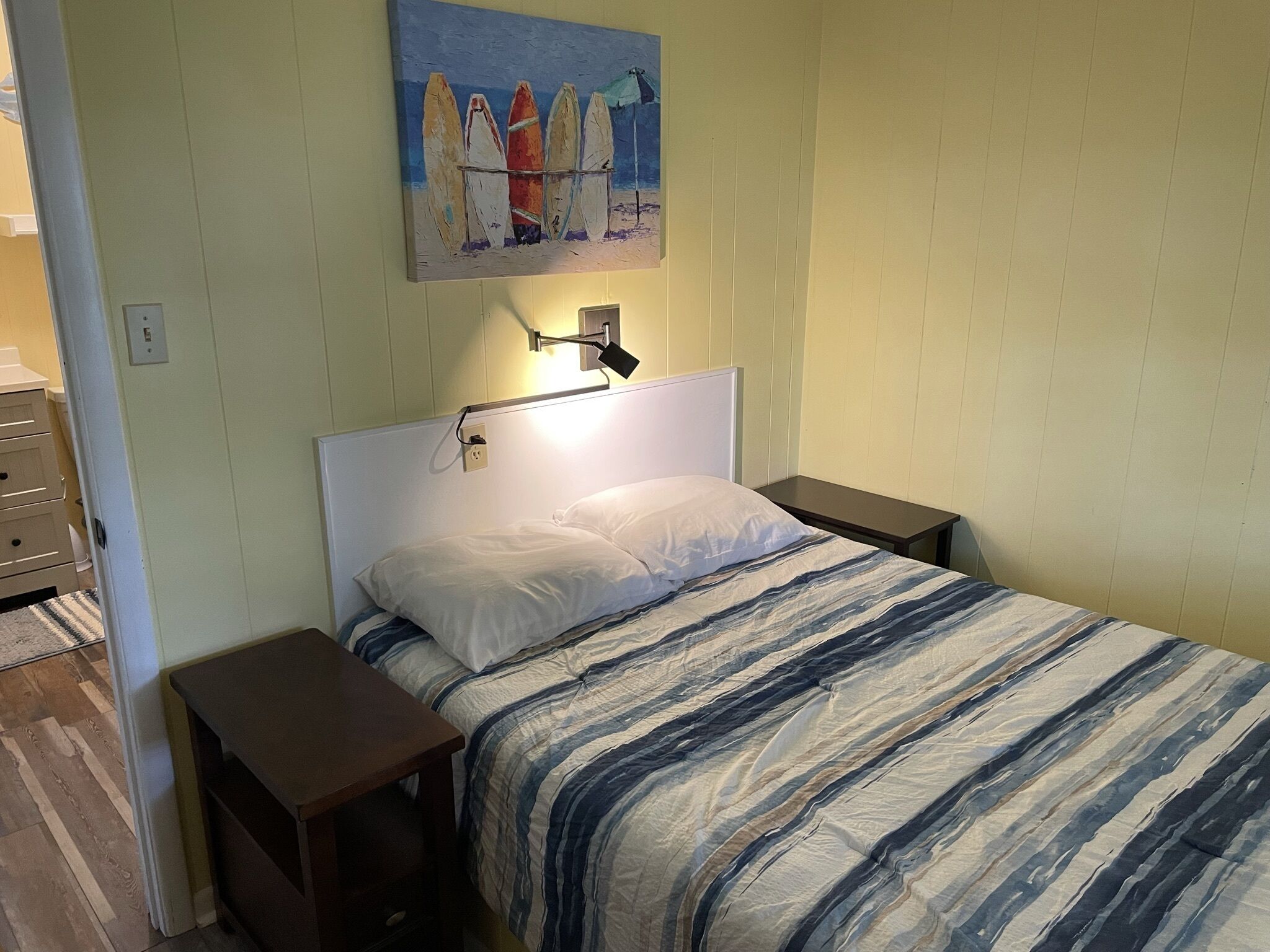 Een slaapkamer met een bed, een nachtkastje, een wandlamp en een schilderij van surfplanken.