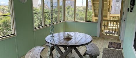 A covered outdoor dining area with a wooden table and benches, large windows, and a view of the neighborhood.