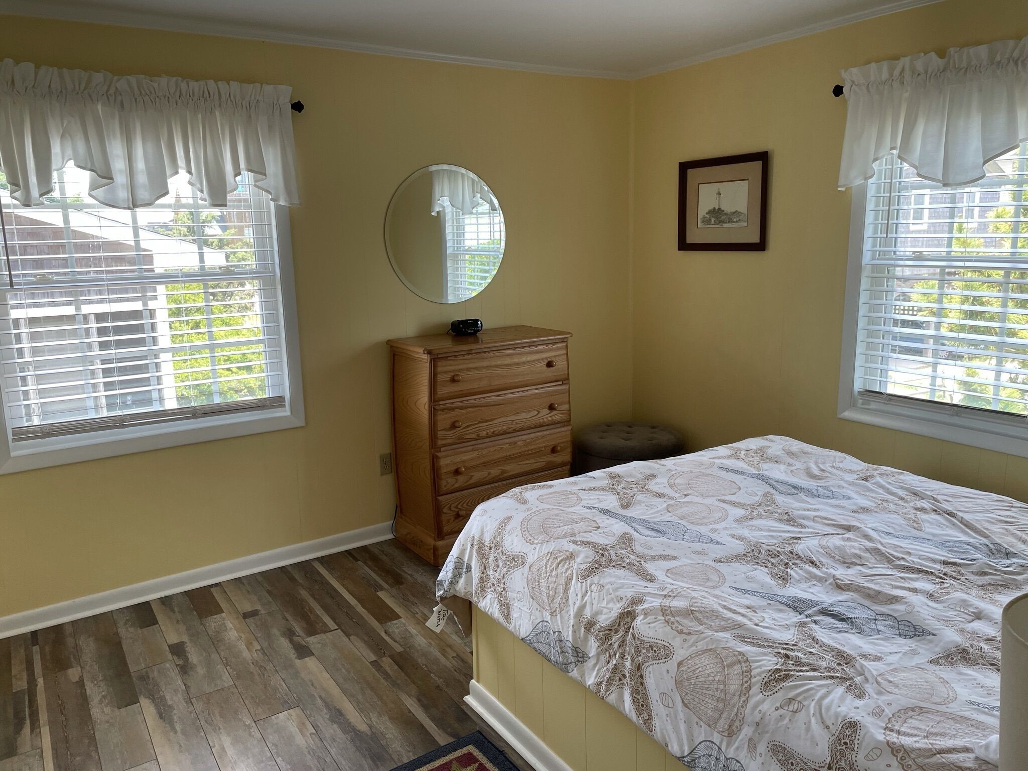 A bedroom with a bed, a dresser, a round mirror, and two windows with curtains.