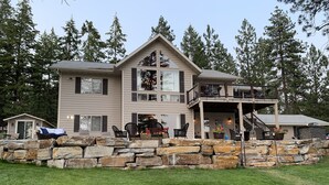 A large, two-story house with a stone retaining wall in front, a covered porch with outdoor seating, and a staircase leading to a second-floor balcony.