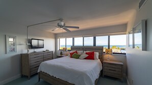 A bedroom with a bed, a ceiling fan, a TV, a dresser, and a window with a view.
