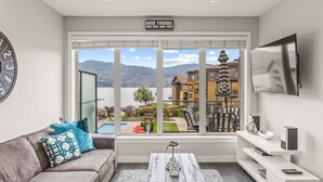 A modern living room with a large window offering a view of a lake and buildings. -  Luxury Beachfront Penthouse Suite. Approved for short-term rentals. (West Kelowna)