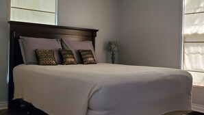 A bedroom with a bed, a nightstand, a lamp, and two windows with blinds.