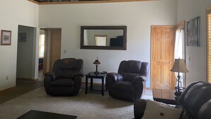 A spacious living room with two leather armchairs, a wooden coffee table, and a chandelier. A large mirror hangs on the wall, and there is a wooden door and a window with curtains.