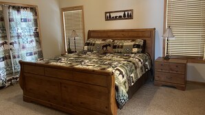 A bedroom with a wooden bed, two bedside tables, a nightstand, a window with blinds, and a curtain with a nature-themed pattern.