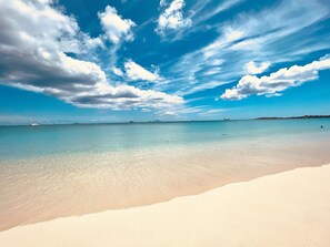 Une plage à l’eau turquoise et limpide, un rivage sablonneux et quelques bateaux visibles au loin.