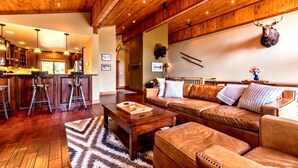 A spacious living area with a wooden ceiling, a leather sofa, a coffee table, and a kitchen in the background.