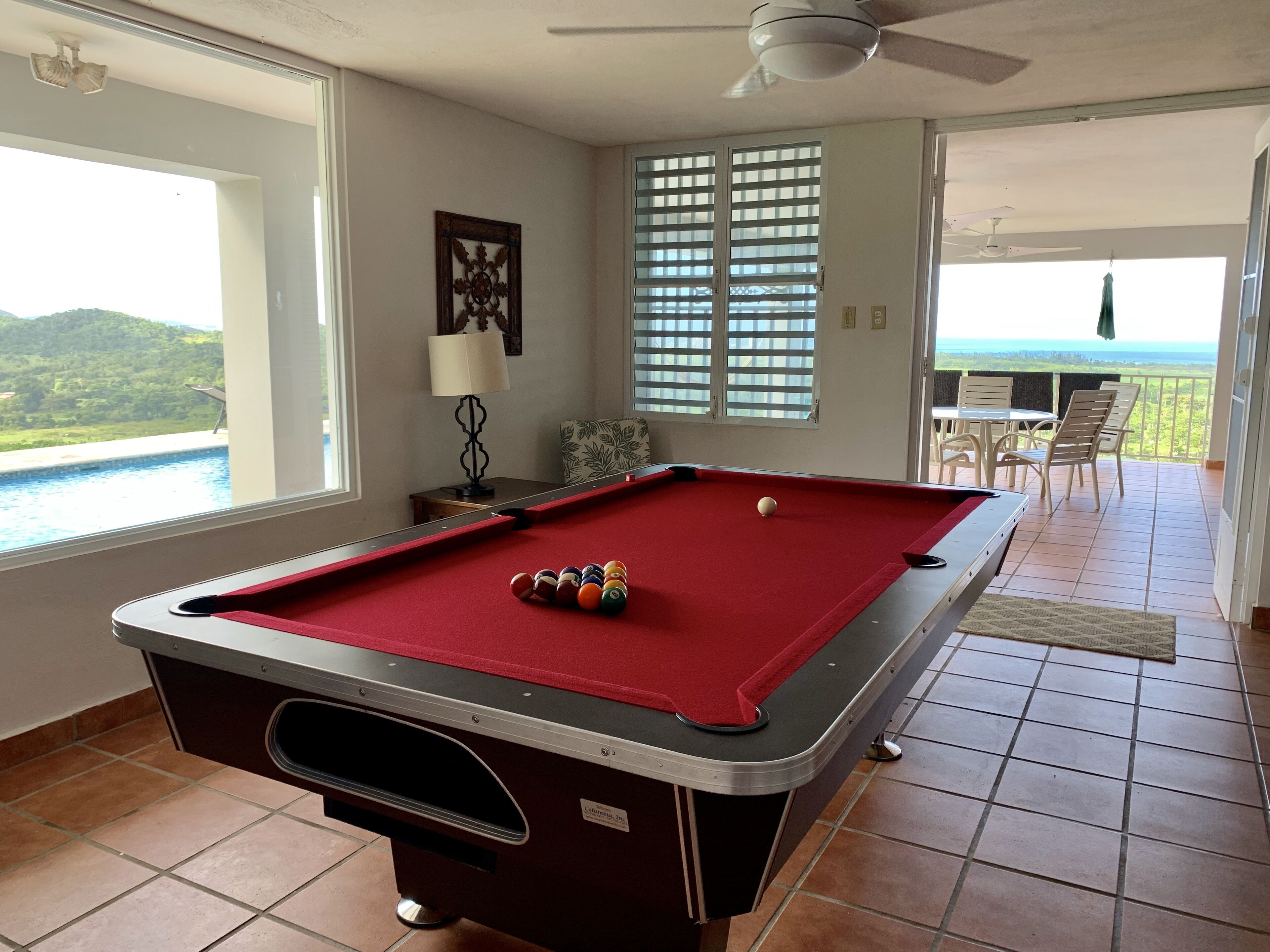 Una mesa de billar con bolas, una lámpara y vista a una piscina.