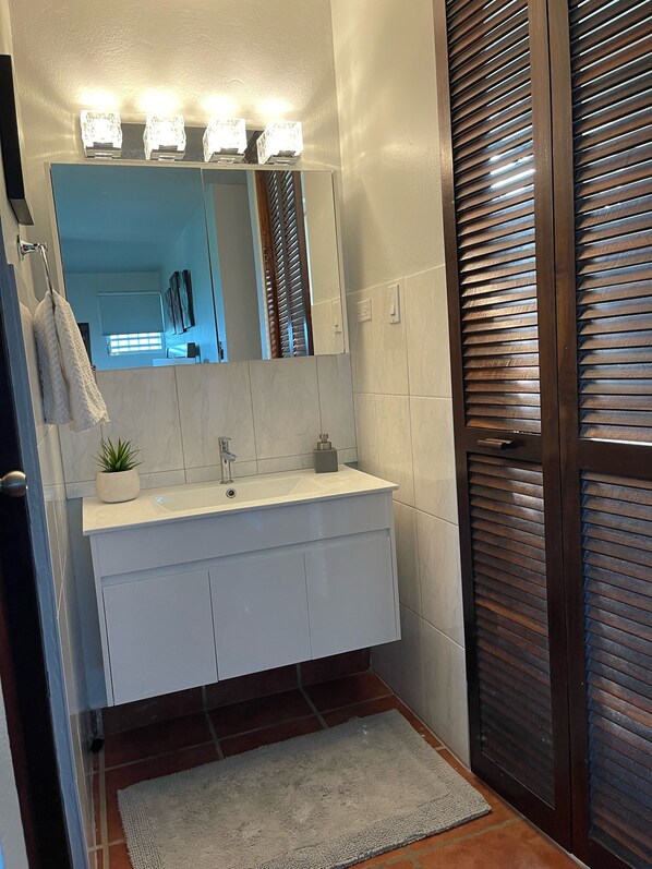 A bathroom with a white vanity, a mirror with lights, a towel rack, and a wooden door with horizontal slats.