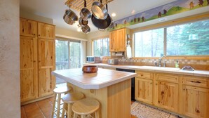 Una cocina con armarios de madera, una isla central con taburetes, un ventanal con vistas a árboles y ollas colgantes.