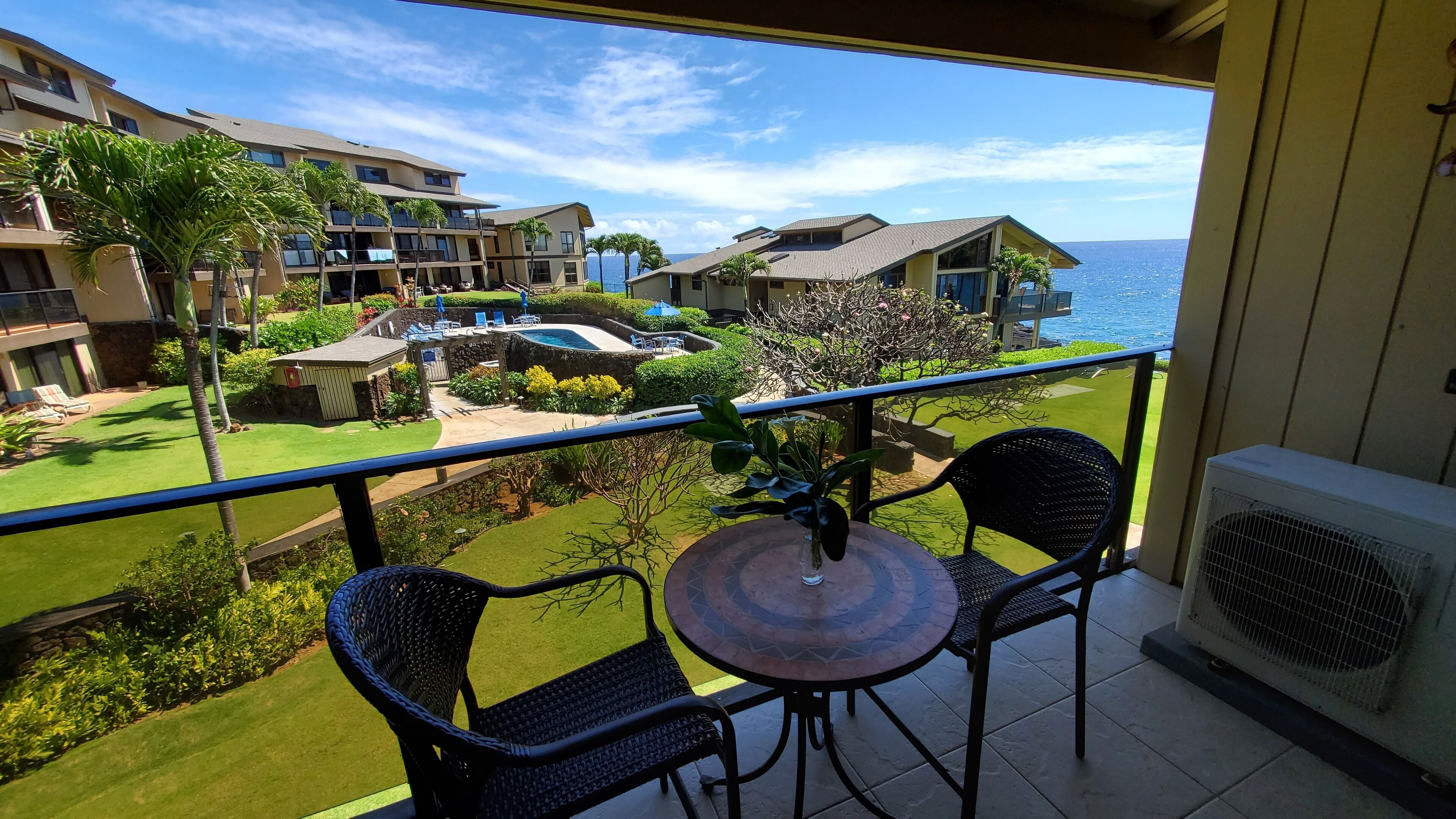 A balcony with two wicker chairs and a small table, overlooking a well-maintained lawn and a pool area with lounge chairs.
