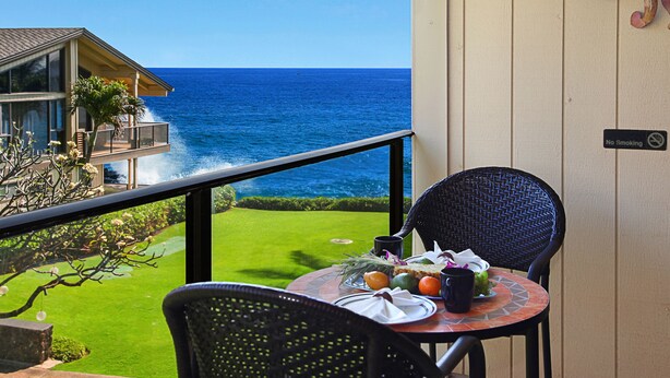 A balcony with a dining table set for two, overlooking a clear blue ocean and a well-maintained lawn. - BEAUTIFUL POIPU OCEAN VIEW 2 BRD 2.5 BATH Condo with A/C 
Great Fall Rates!
 (Koloa)