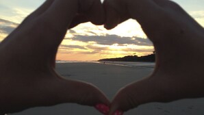 Mãos formando um coração com uma praia ao pôr do sol ao fundo.
