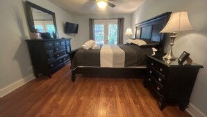 Quarto com cama, cômoda, mesa de cabeceira, ventoinha no teto e televisão na parede.