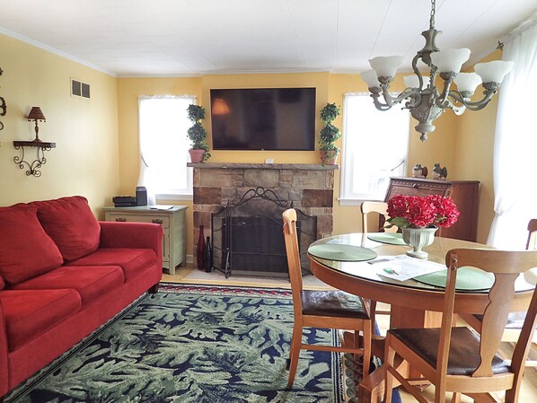 A living room with a red sofa, a fireplace, a television mounted above it, and a dining area with a table and chairs. - Charming 2B/1B Cottage, Private Patio, Downtown, OffStreet Parking, Dog Friendly (Saugatuck)