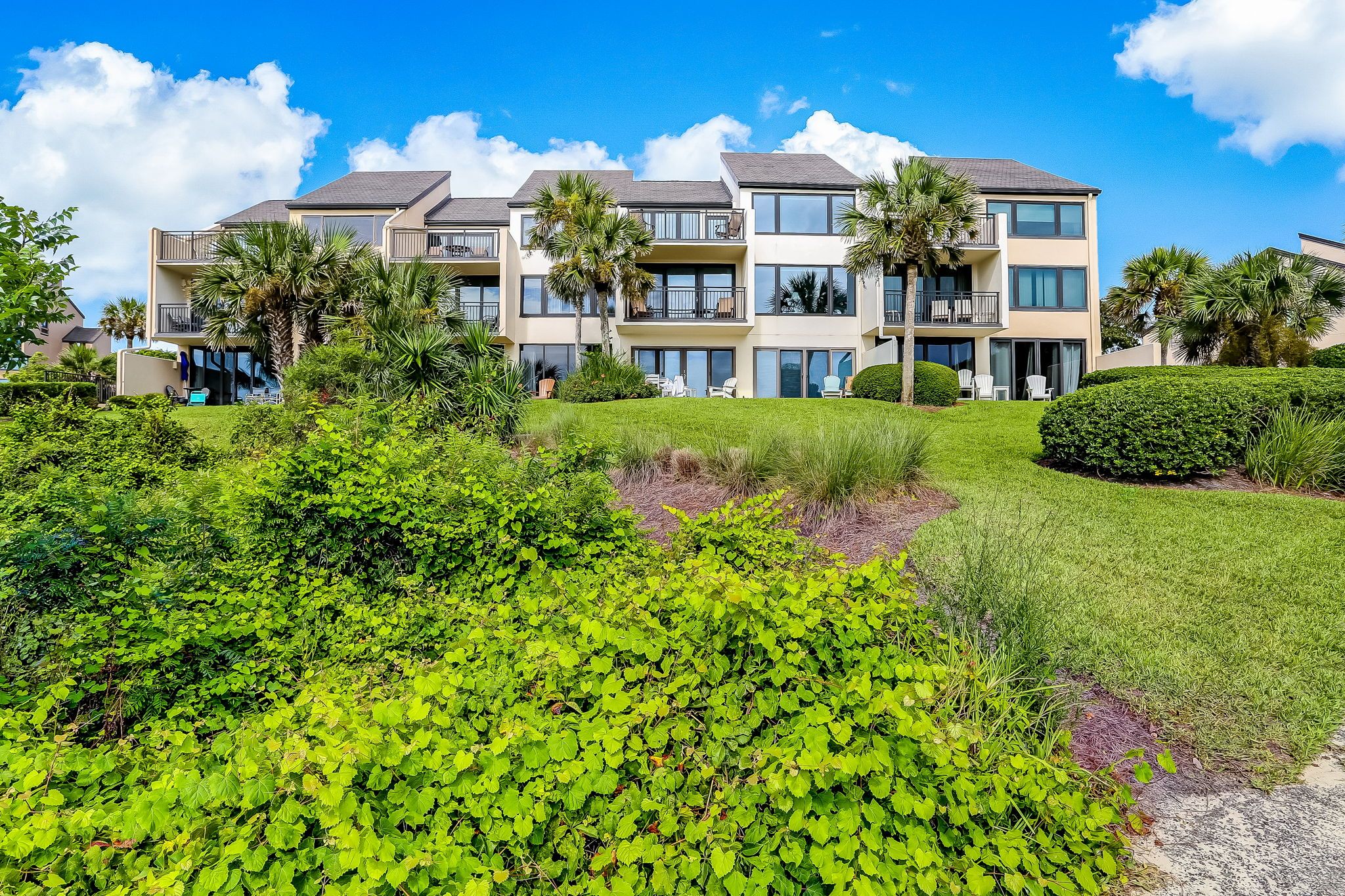 A multi-story residential building with balconies, surrounded by lush greenery and palm trees.