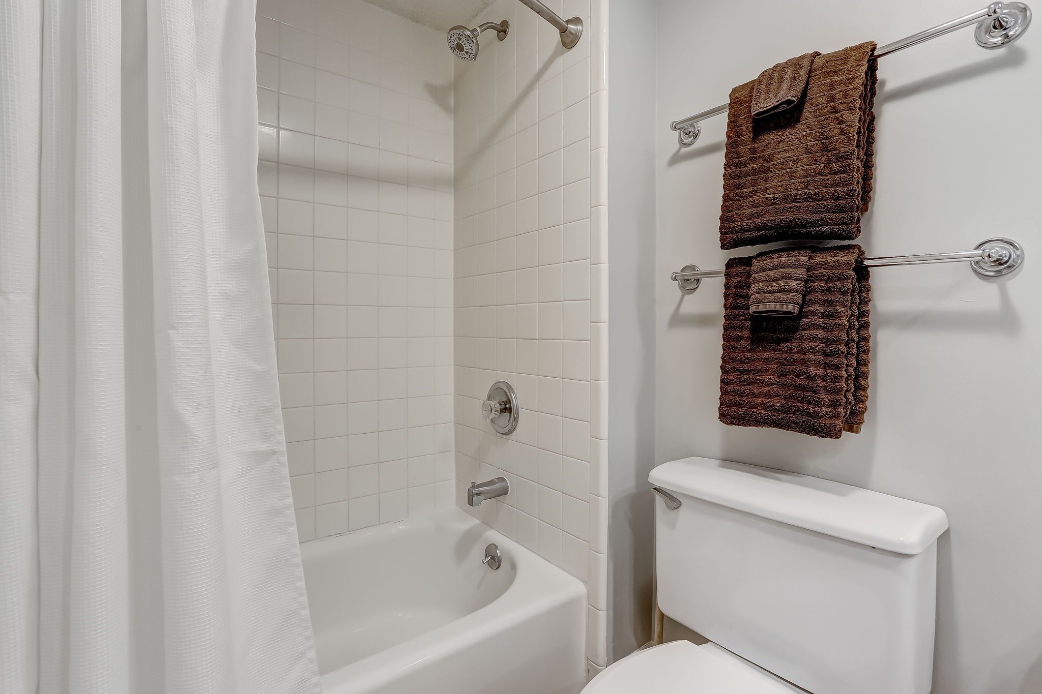Un baño con una bañera blanca, baldosas blancas y dos toallas marrones colgadas en un perchero.