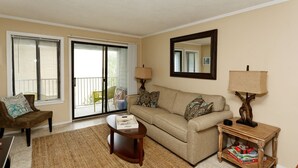 A living room with a sofa, armchair, coffee table, and a wooden side table with a lamp. There is a sliding glass door leading to a balcony. - Beach Resort, Walk to the Ocean, Free Secure Wi-Fi in Building 1 (Hilton Head Island)