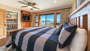 A bedroom with a large bed, a ceiling fan, a wooden dresser, and a view of the water and mountains.