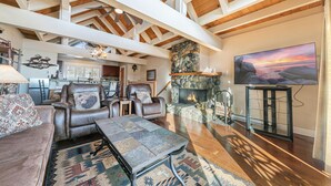 A spacious living room with leather sofas, a stone fireplace, a television, and a high ceiling with exposed beams.