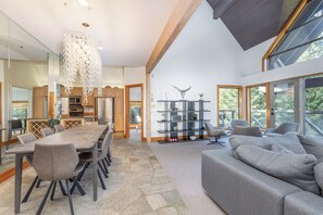 A spacious living area with a large gray sofa, a dining table with chairs, a decorative chandelier, and a bookshelf with decorative items.