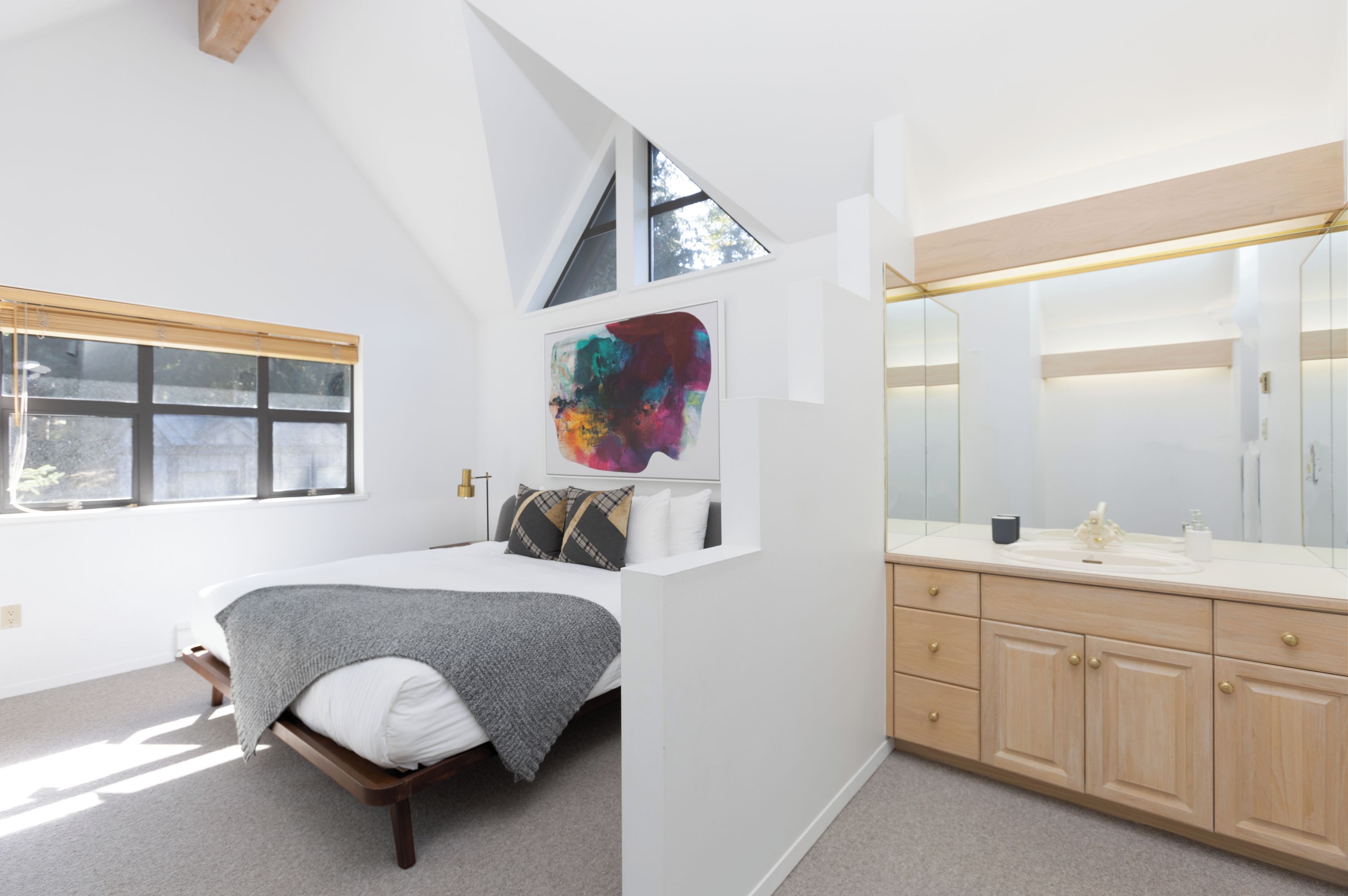 Un dormitorio moderno con cama, un ventanal amplio, un baño con espejo y lavabo, y un cuadro abstracto colorido en la pared.