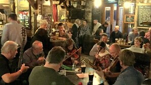 Un bar concurrido donde gente está sentada y de pie, tocando instrumentos musicales, y un cartel de menú a la vista al fondo.