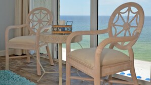 A room with two wooden chairs and a small table, a blue rug, and a view of the ocean through large windows.