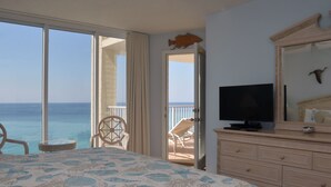 A bedroom with a large bed, a TV, a dresser, and a balcony with a view of the ocean.