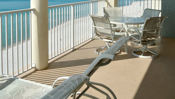 A balcony with white railings, a table, and two lounge chairs facing a beach.
