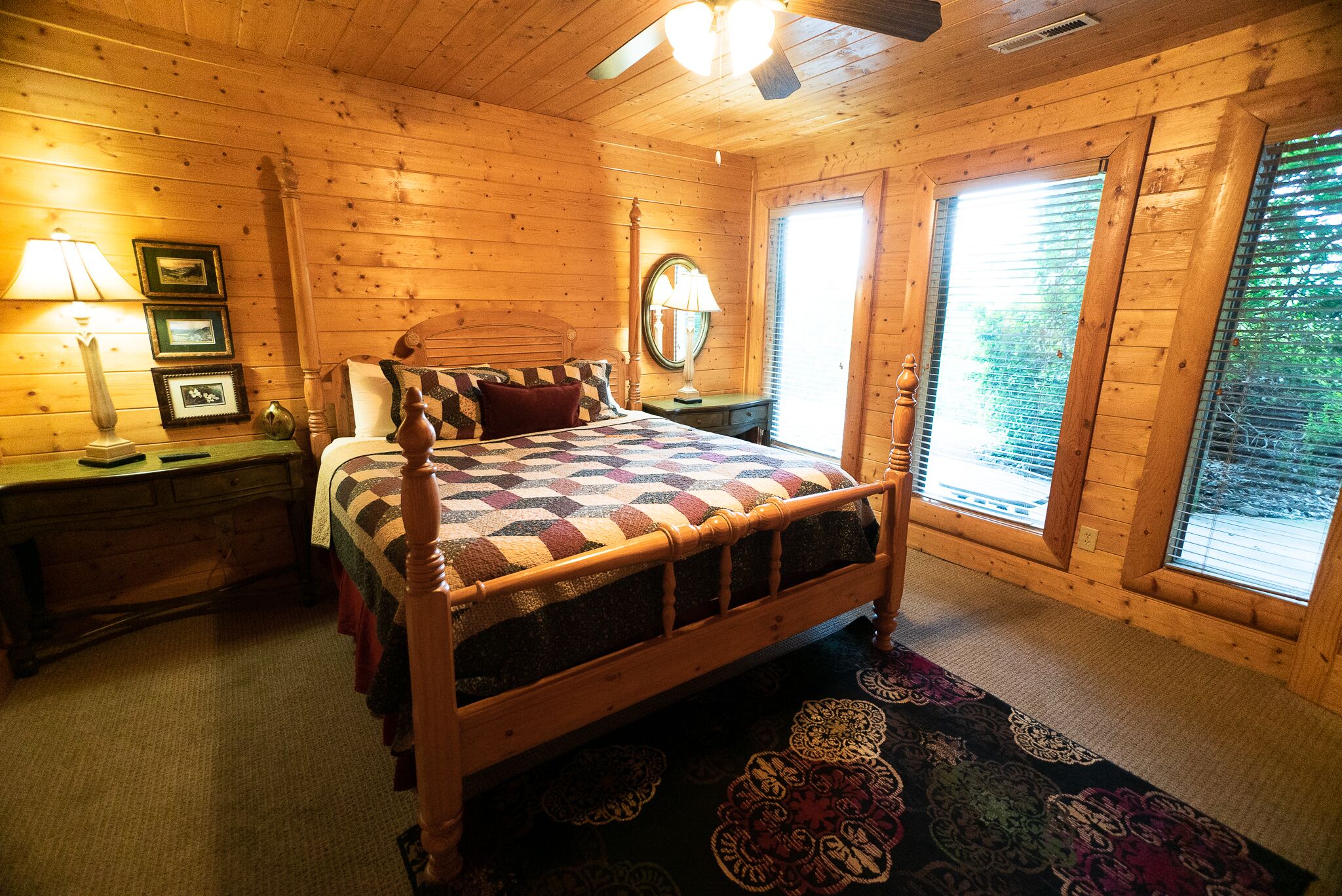 A wooden cabin bedroom with a bed, bedside tables, a mirror, and large windows.