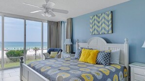 Un dormitorio con cama, ventilador de techo, mesita de noche, lámpara, vista a la playa y un cuadro decorativo con diseño en patrón de chevron.