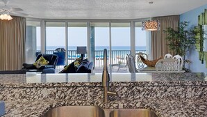 Una sala de estar moderna con un gran ventanal que da a una playa, un ventilador de techo y un comedor con sillas blancas.