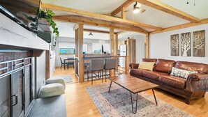 A spacious living area with a brown leather sofa, a coffee table, and a dining table with chairs. There is a kitchen area in the background with wooden beams and a hanging plant.