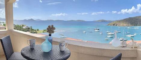 A balcony with wicker chairs and a table, overlooking a marina with boats and a clear blue sea.