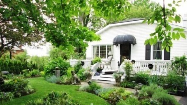A white house with a covered porch, surrounded by a well-maintained garden with various plants and a lawn. - Alnwick Cottage, quaint and comfortable (Niagara on the Lake)
