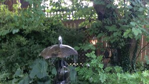 A garden with a fountain featuring a statue of a figure holding a shield, surrounded by lush greenery and a wooden fence in the background. - Alnwick Cottage, quaint and comfortable (Niagara on the Lake)
