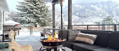 A cozy outdoor seating area with a wood-burning stove, a sofa, and a view of snow-covered mountains.