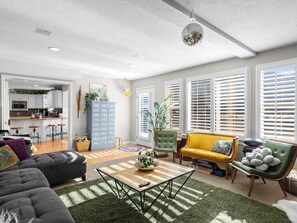 Ein geräumiges Wohnzimmer mit Sofa, Sessel und Couchtisch, im Hintergrund eine Küche und große Fenster mit Jalousien.