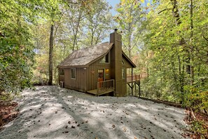 A rustic cabin with a chimney, a porch, and a gravel driveway surrounded by trees.