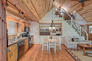 A rustic cabin interior with a wooden ceiling, a dining area with white chairs, a sofa, and a staircase with a natural wood railing.
